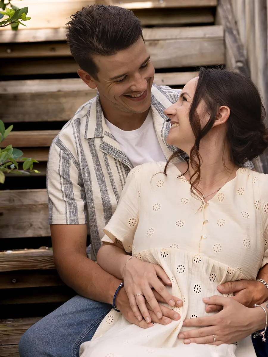Un couple complice se regardant avec tendresse, les mains posées sur le ventre de la femme enceinte, assis sur des escaliers en bois