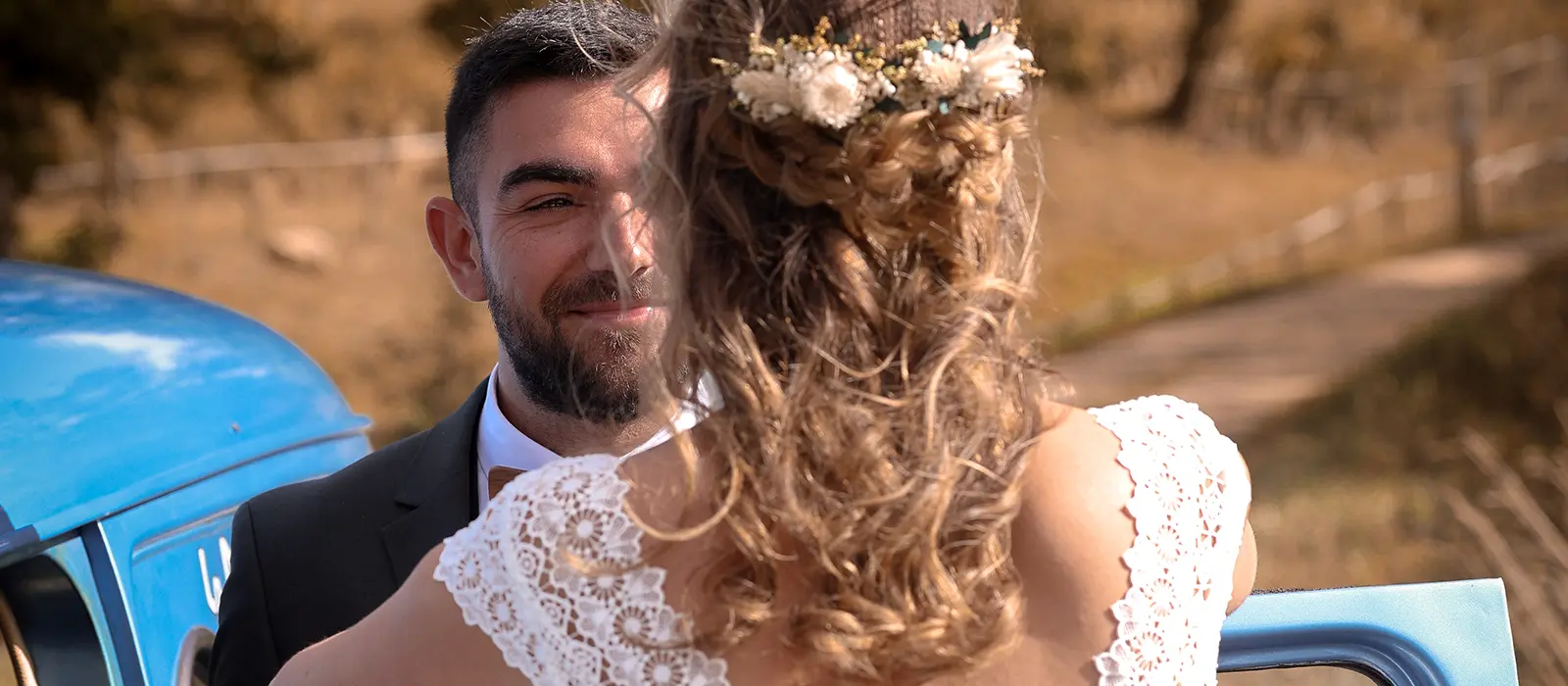 Le marié souriant à la mariée vue de dos avec sa robe en dentelle, appuyée sur une voiture bleue vintage