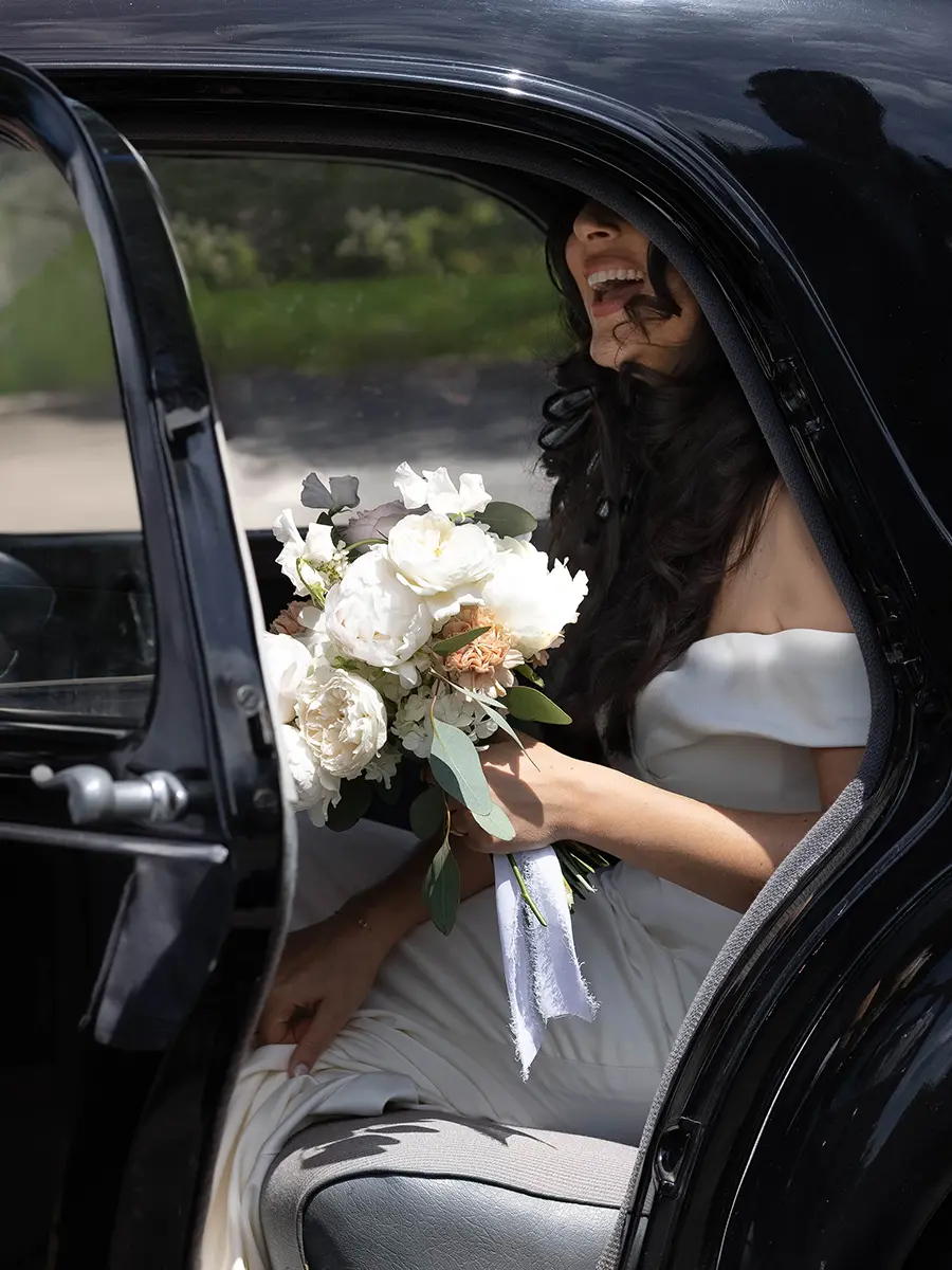 Une mariée souriante tenant son bouquet de fleurs blanches à l'arrière d'une voiture ancienne noire